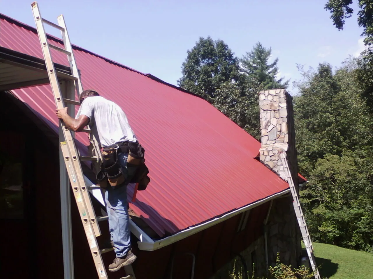 Roofer on a ladder beside a red metal roof and stone chimney — Soffit & Fascia Repair by Roofing Brothers in Francisco